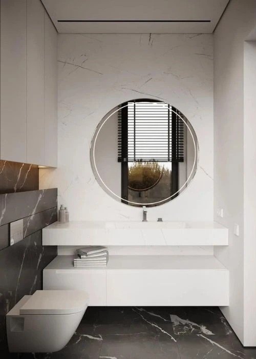 Baño con paredes de mármol blanco, espejo circular y suelo oscuro